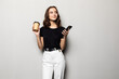 © dianagrytsku - Portrait of a young smiling girl in shirt texting message on mobile phone and holding cup of coffee to go over gray background