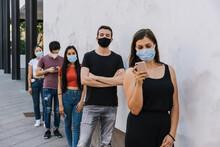 Group Of Young People At Safety Distance Near A White Wall In Queue In Protection By Coronavirus, Covid-19 With Face Mask While Using The Device - Multiracial Group Of Friends - Concept Of Security