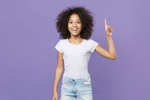 Excited Little African American Kid Girl 12-13 Years Old In White T-shirt Isolated On Violet Wall Background Studio Portrait. Childhood Lifestyle Concept. Holding Index Finger Up With Great New Idea.