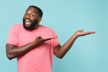 Cheerful young african american man guy in casual pink t-shirt isolated on blue background studio portrait. People emotions lifestyle concept. Mock up copy space. Pointing index finger hand aside.