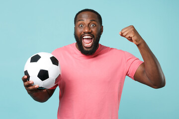Wall Mural - Excited african american man guy football fan in pink t-shirt isolated on blue wall background. Sport family leisure lifestyle concept. Cheer up support favorite team with soccer ball, clenching fist.