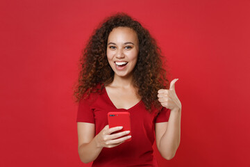 Wall Mural - Funny young african american girl in casual t-shirt posing isolated on red wall background studio portrait. People lifestyle concept. Mock up copy space. Using mobile cell phone, showing thumb up.
