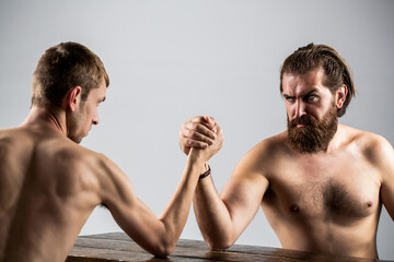 Wall Mural - Arm wrestling. Heavily muscled bearded man arm wrestling a puny weak man. Arms wrestling thin hand, big strong arm in studio. Two man's hands clasped arm wrestling, strong and weak, unequal match