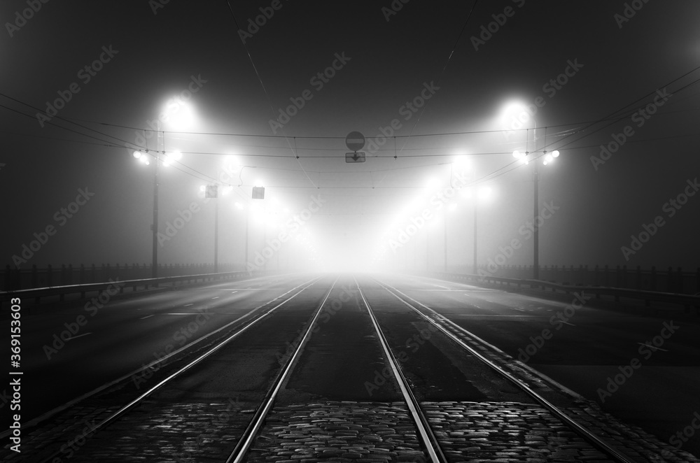 Tramway track and asphalt road (highway) through the illuminated empty Stone bridge in a thick fog at night. Lanterns close-up. Daugava river, Riga, Latvia. Concept image, black and white, monochrome - obrazy, fototapety, plakaty 