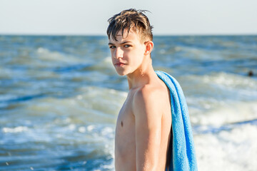 Teenager with a blue towel on his shoulder against the background of the sea. Splashes from the sea wave. Summer vacation at the sea. To swim in the sea. Summer tourism. Waves in the ocean.