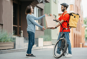 Courier with bicycle delivered shopping bag to client. Smiling girl takes bag from man in helmet with backpack