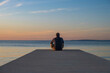 Man in hoodie and short cargo pants sitting on the edge od dock and meditating. Sea horizon, island od Pag, Adriatic sea, Croatia
