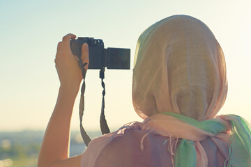 Wall Mural - Arab Woman Photographer in a scarf taking picture using Camera on the sunset background. Halal travel concept