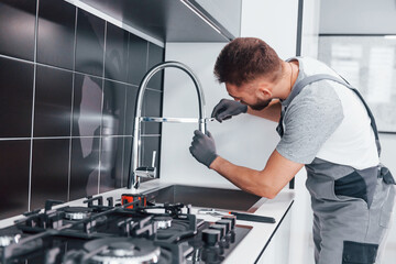 young professional plumber in grey uniform fixing water tap on the kitchen