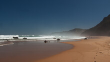 Ocean Coast With Footprints On Sandy Beach, Black Rocks, Blue Sky And Huge Waves. Portugal, West Coast