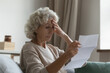 Disappointed elderly woman sitting on couch in living room feels frustrated by bad news, reading paper postal correspondence, bank notice about debt, notification of eviction, negative letter concept