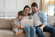 © fizkes - Happy young woman holding computer on laps, watching funny comedian movie video or shopping online with loving husband and adorable little child son, relaxing together on couch in living room.