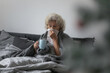 Senior woman sit on bed in bedroom wrapped in warm knitted plaid holds cup with antipyretic healing beverage, blow runny nose use paper handkerchief. Got hay fever, rhinitis, allergy symptoms concept