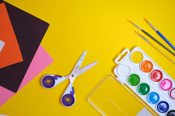 back to school concept, paintbrush, watercolor paints, scissors and velvet colored paper on yellow background, copy space, top view