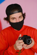 © Oleg Samoylov - Male photographer. A young man of 25-30 years old in a black protective mask and in a red sweatshirt holds a mirrorless camera in his hands.