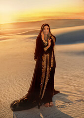 Beautiful mysterious arab woman in black muslim abaya dress. head is twisted with silk handkerchief chain hides face. Fantasy Girl walks in desert, art background sand golden sunset. Queen Concubine