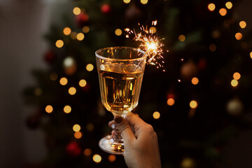 Wall Mural - Burning firework in champagne glass in hand on background of golden bokeh lights in festive room. Happy New Year. Hand holding drink with sparkler at christmas tree. Creative aesthetic moment