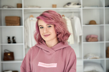 Smiling hipster gen z teen girl fashion designer, stylist with pink hair piercing wearing hoodie looking at camera in front of modern clothes wardrobe closet, face head shot close up portrait.