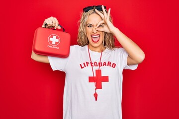 Beautiful blonde lifeguard woman wearing t-shirt with red cross and whistle holding first aid kit smiling happy doing ok sign with hand on eye looking through fingers