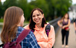 Happy students talking after studies. Cheerful young woman with backpack smiling and talking with female friend while standing on bridge after university studies