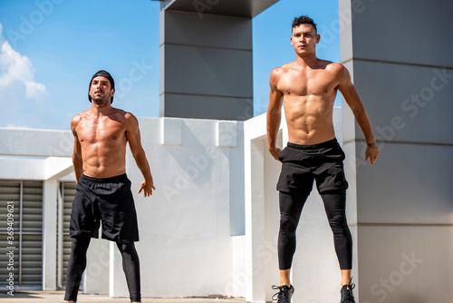 Shirtless handsome athletic men doing squat jump workout outdoors on rooftop, no equipment home exercise concept