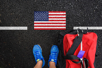 a man with a shoes and travel bag is standing on asphalt next to flag of United States and border