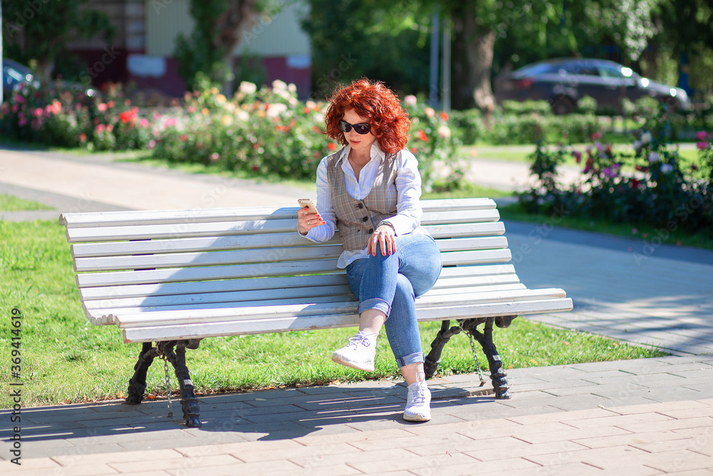 Red-haired curly girl in a shirt and jeans sits on a bench in the park ...