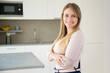 © Mangostar - Cheerful attractive fair haired young woman posing with arms folded in kitchen, looking at camera and smiling. Medium shot, copy space. Woman at home or female portrait concept
