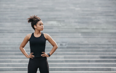 Portrait of athlete at start of training. Serious girl with beautiful body with wireless headphones and smart watch stands on city stairs background