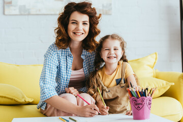 Wall Mural - happy nanny and kid smiling at camera while drawing together