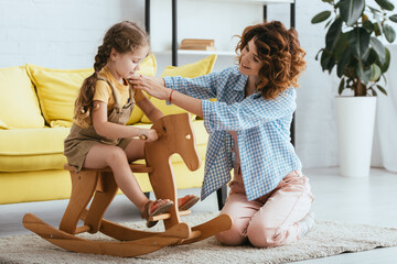 Wall Mural - smiling nanny touching adorable kid riding rocking horse at home