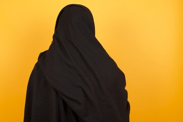 the back view of a middle aged muslim woman wearing black hijab over yellow background studio shoot.