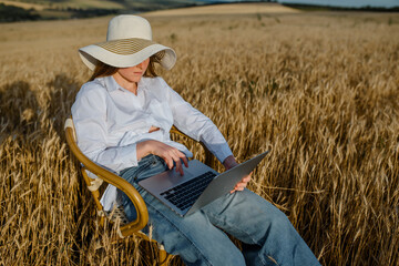 Young woman freelancer is working on a computer outdoors in the wheat field. She is sitting on a vintage chair and enjoying the ability to work from anywhere