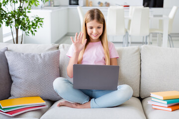 Wall Mural - Full length photo of little kid girl sit cozy couch legs crossed have online lesson conversation greet teacher wave hand use laptop study remote in house indoors