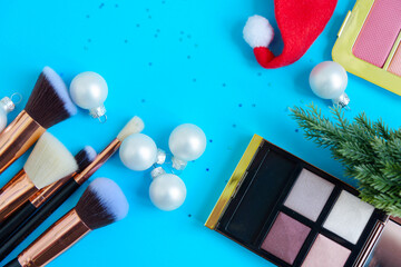 Wall Mural - creative Christmas cosmetics flat lay - premium makeup brushes, blush palette, eyeshadow palette, Christmas balls, Christmas tree and Santa hat on a colored blue background