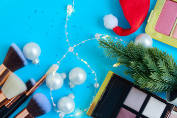 Wall Mural - creative Christmas cosmetics flat lay - premium makeup brushes, blush palette, eyeshadow palette, Christmas balls, Christmas tree and Santa hat on a colored blue background