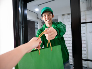 Asian young delivery man in green uniform jacket smile and send shopping paper bag to customer who purchased order online look like door to door service with quality at home office