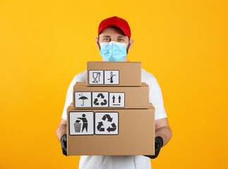 Courier in mask holding cardboard boxes with different packaging symbols on yellow background. Parcel delivery