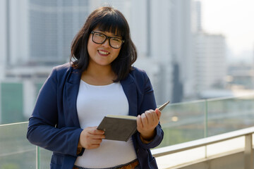 Happy young beautiful overweight Asian businesswoman against view of the city