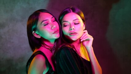 Wall Mural - Portrait of two young flirting cute Asian women playfully posing for camera on the pink and blue neon lights - video in slow motion