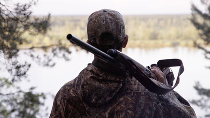 View from back of hunter in camouflage puts hunting rifle on his shoulder and looks at river from forest. Outdoor activity concept. Summer hiking in wilderness area.