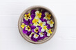Pansies in a wooden bowl. Edible flowers, field pansies, violets on a white wooden table. Concept of edible flowers. Cuisine ingredient, condiment.