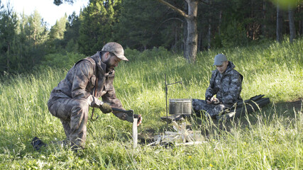 Man in camouflage clothes is chopping wood with axe and throwing it into bonfire. Tourist concept. Hunter checks or cleans hunting rifle. Best friends spend leisure weekend in forest nature background