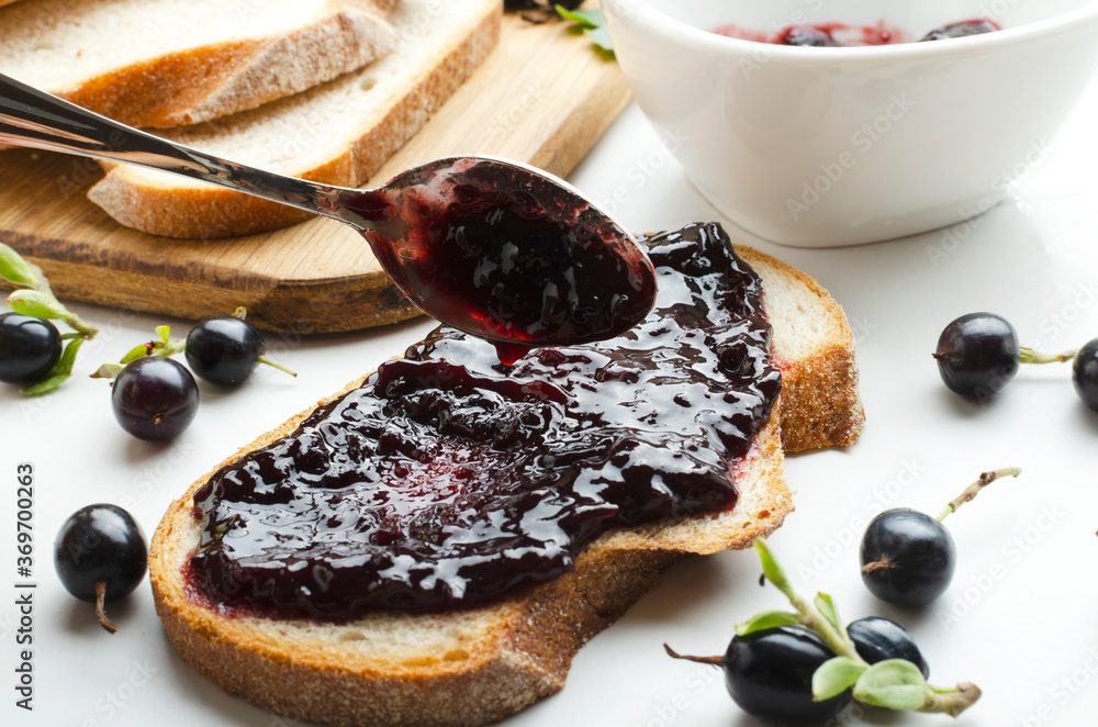 Process of spreading of sweet delicious jam on the slice of bread.Closeup of spoon, blackcurrant jam, berries, bread on the white table.Tasty jam sandwich