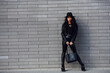 © standret - Beautiful brunette with curly hair and in black clothes holding bag outdoors near wall