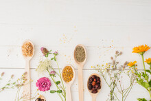 Top View Of Herbs In Spoons And Flowers On White Wooden Background, Naturopathy Concept