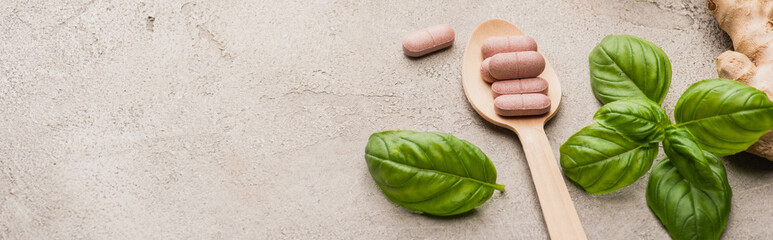 Wall Mural - panoramic shot of green leaves, ginger root and pills in wooden spoon on concrete background, naturopathy concept