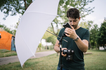 Wall Mural - Male photographer setting flash light while holding camera. Man looking at camera and setting flash light on stand while standing in park.