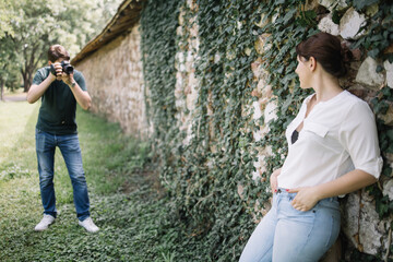 Wall Mural - Photographer shooting woman leaning against wall with ivy. Man with professional camera taking pictures of woman who is posing on old wall.