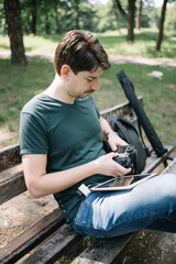 Wall Mural - Side view of man transferring photos from camera outdoor. Male photographer checking images on camera while sitting on bench with tablet in nature.